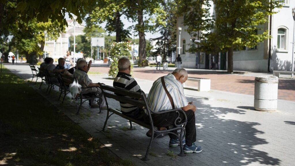 donice z kwiatami, ławki na których siedzą osoby, alejka spacerowa pomiędzy zabudowaniami